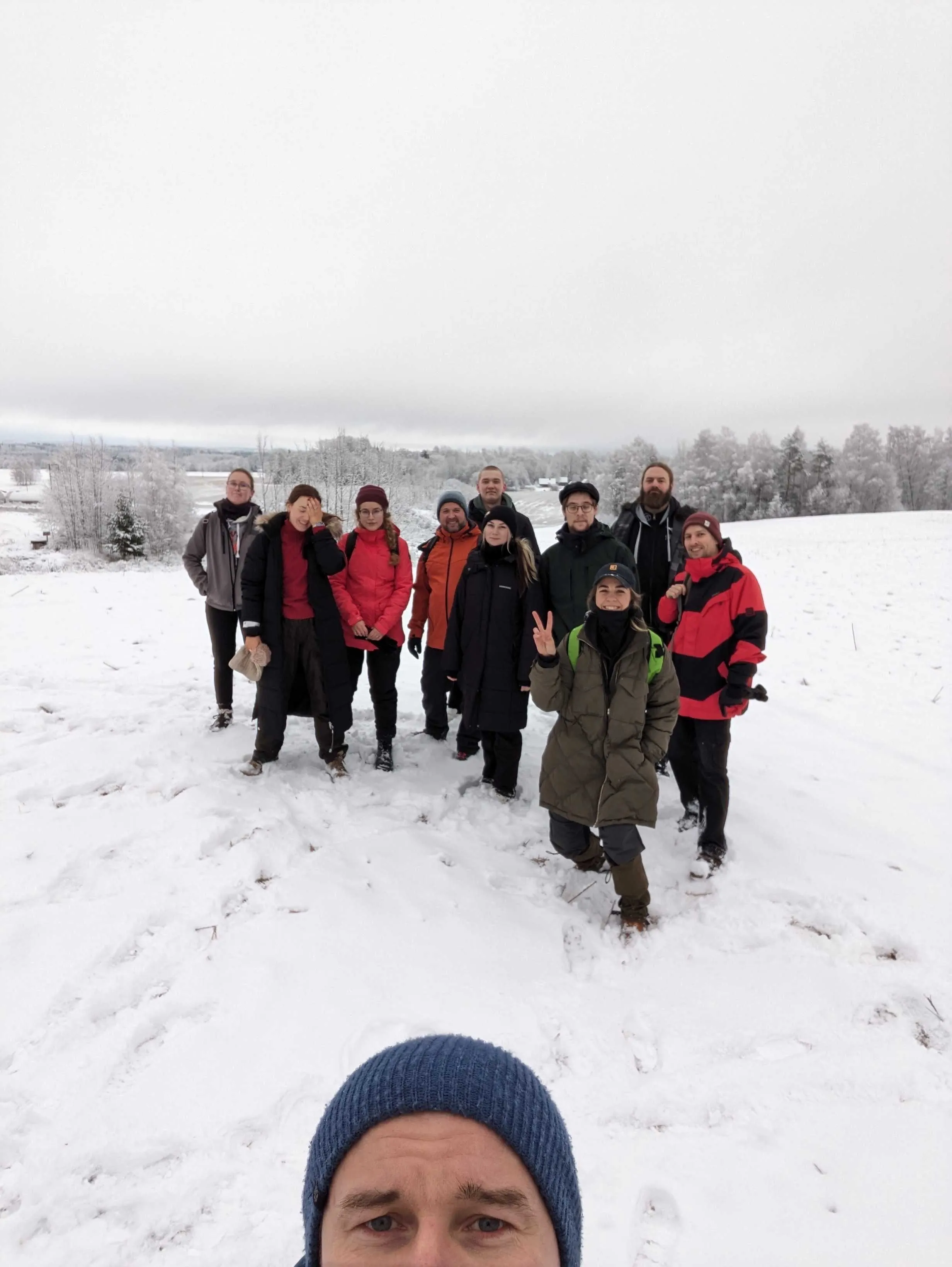 A group of ten people posing in a snowy field, photographed from below by someone in a blue beanie in the foreground.