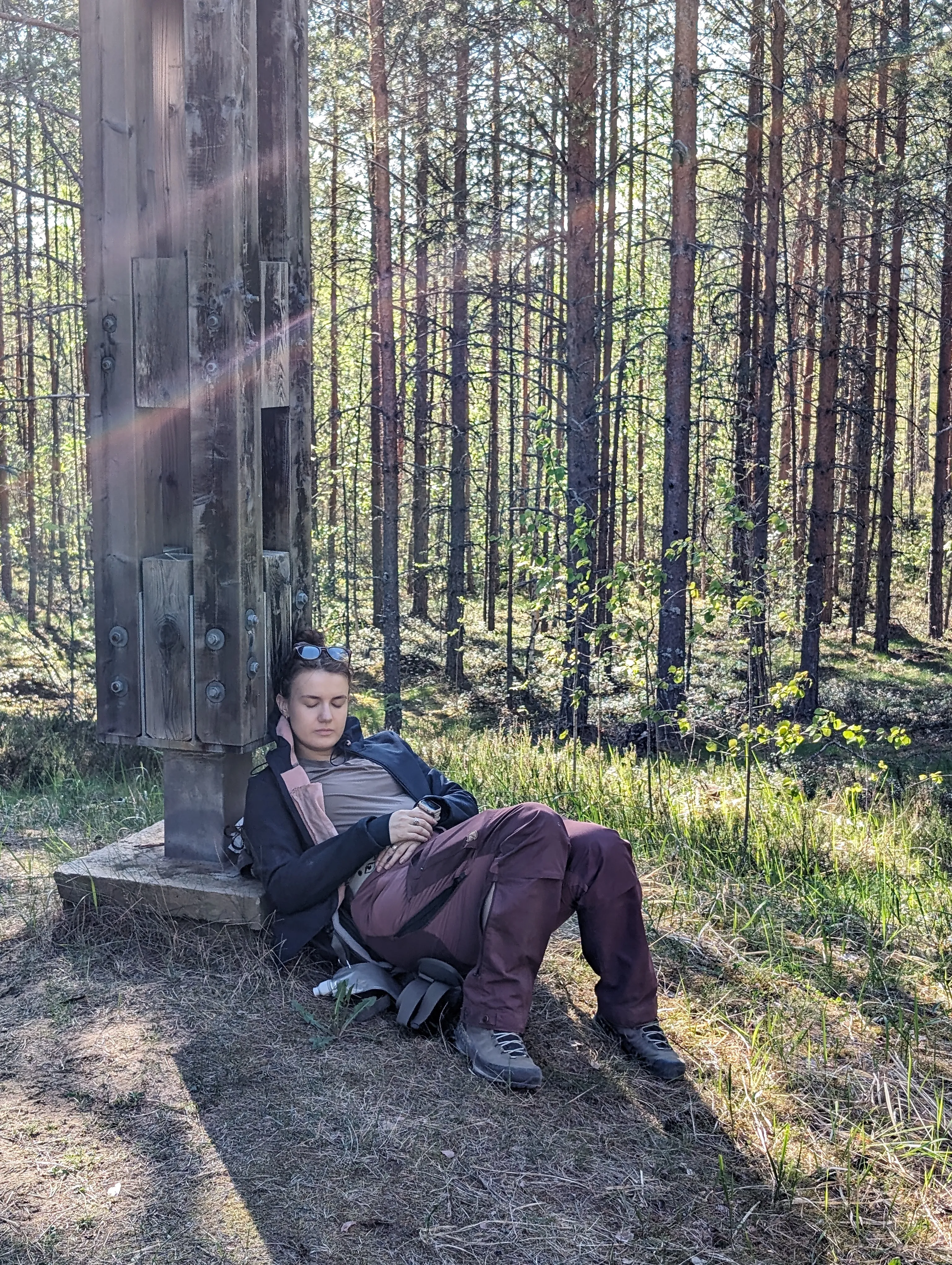A hiker asleep sitting against a tall wooden lookout tower post, backpack still on, in a sunlit pine forest.