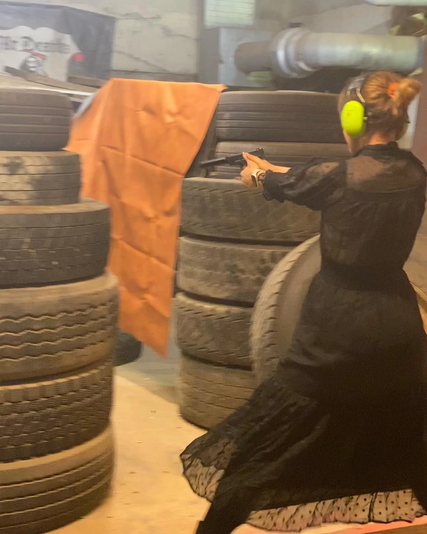 Stella in a black lace dress, firing a handgun at an indoor shooting range stacked with tyres.