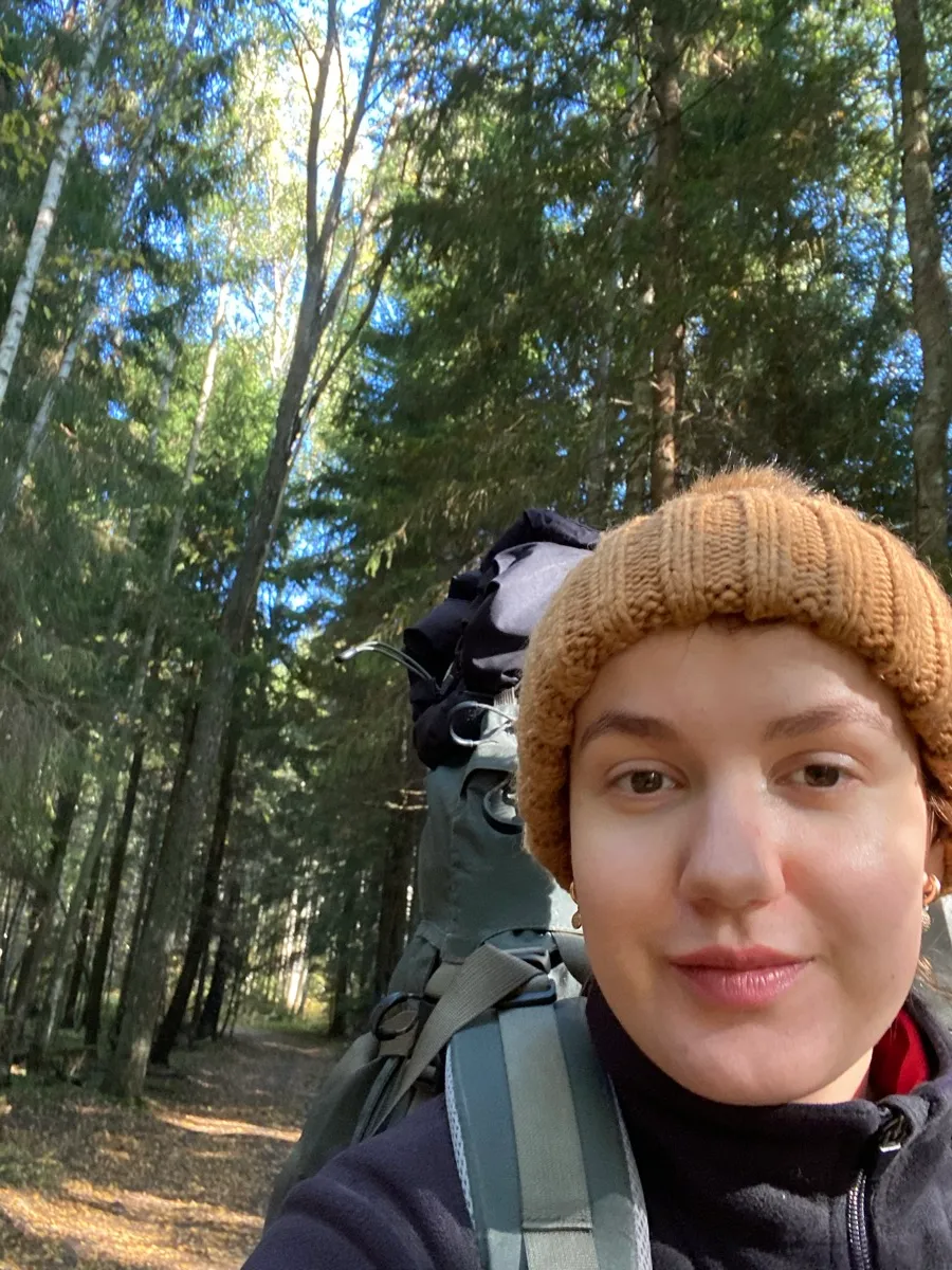 A close-up selfie of a hiker in a knit beanie with a loaded backpack, standing on a sunlit forest trail.