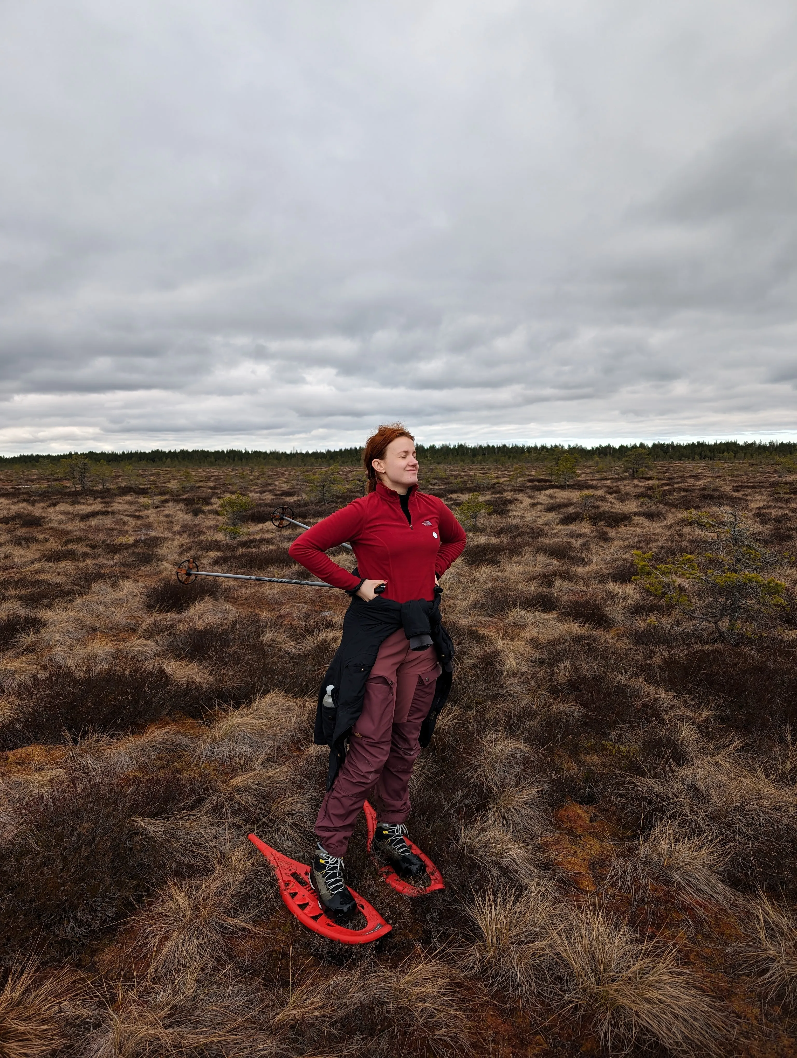 A woman in a red fleece and snowshoes standing on a bog under an overcast sky, ski poles slung over her shoulders.